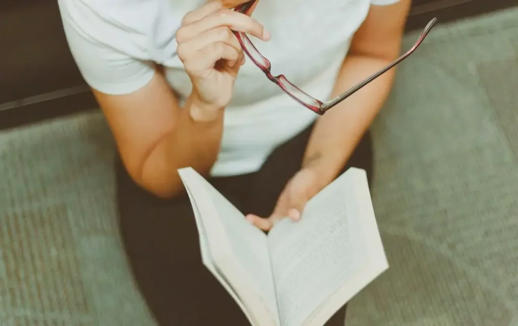 eine Person hält ein aufgeschlagenes Buch und eine Lesebrille